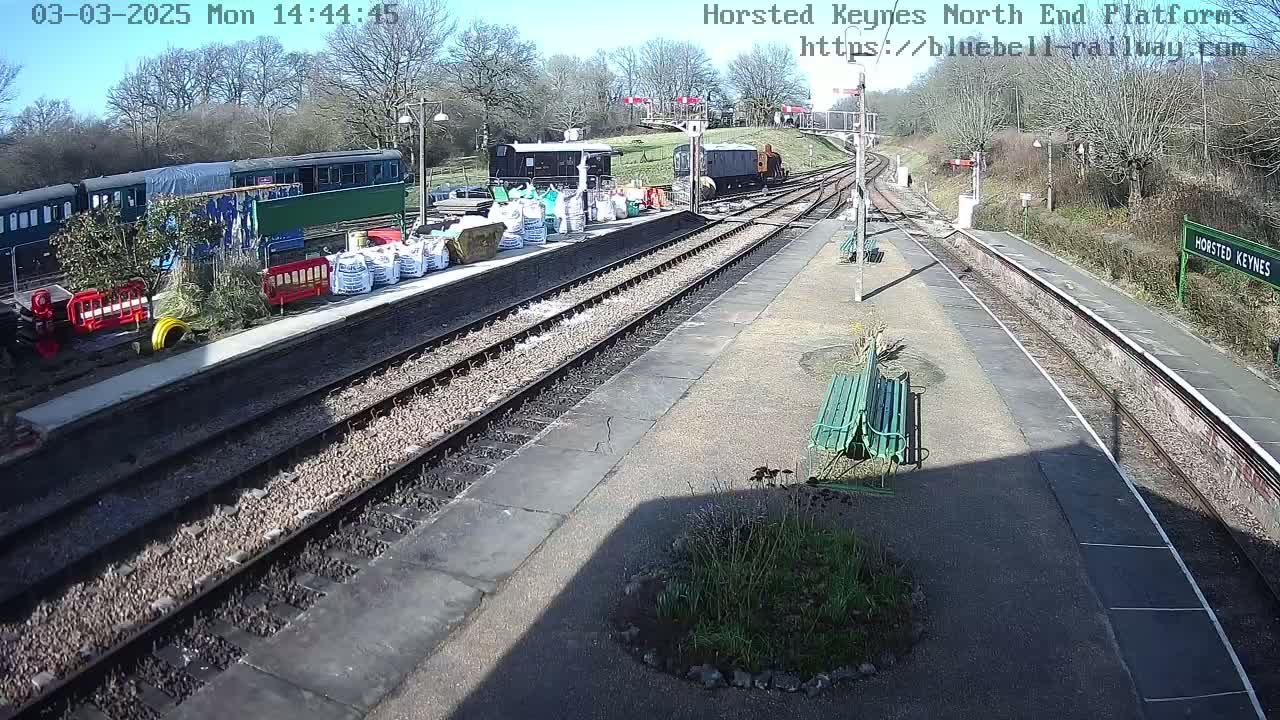 A sunny view of a train station platform with several train cars and some construction materials visible, including bags of what looks like gravel or cement.