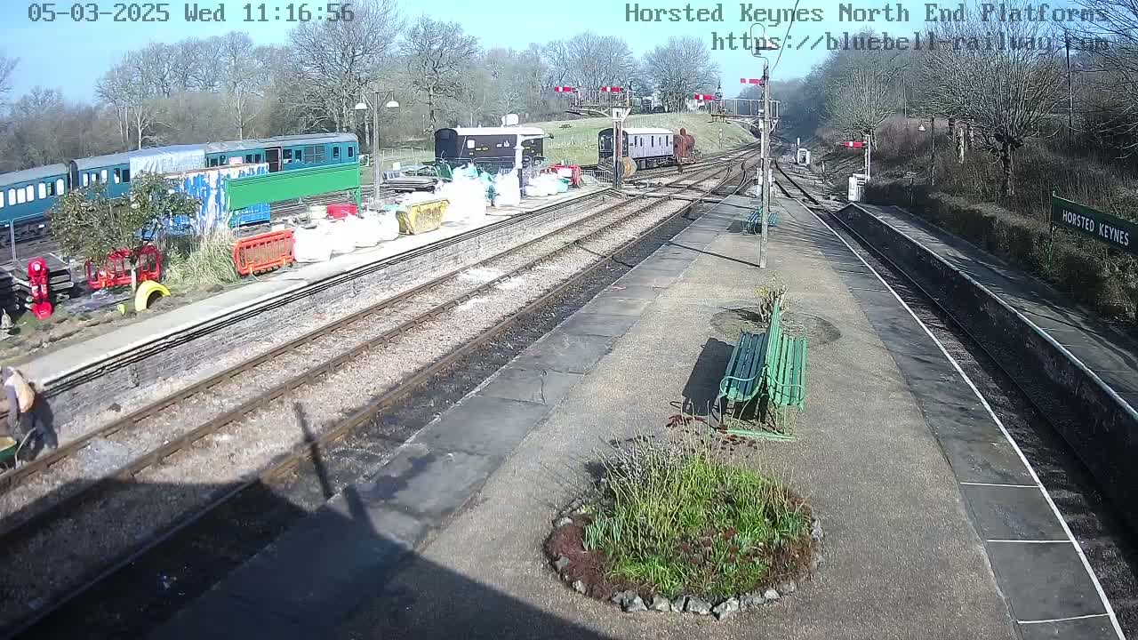 A sunny day at a train station shows several train cars parked near construction materials and equipment, with a few people visible and a couple of benches on the platform.