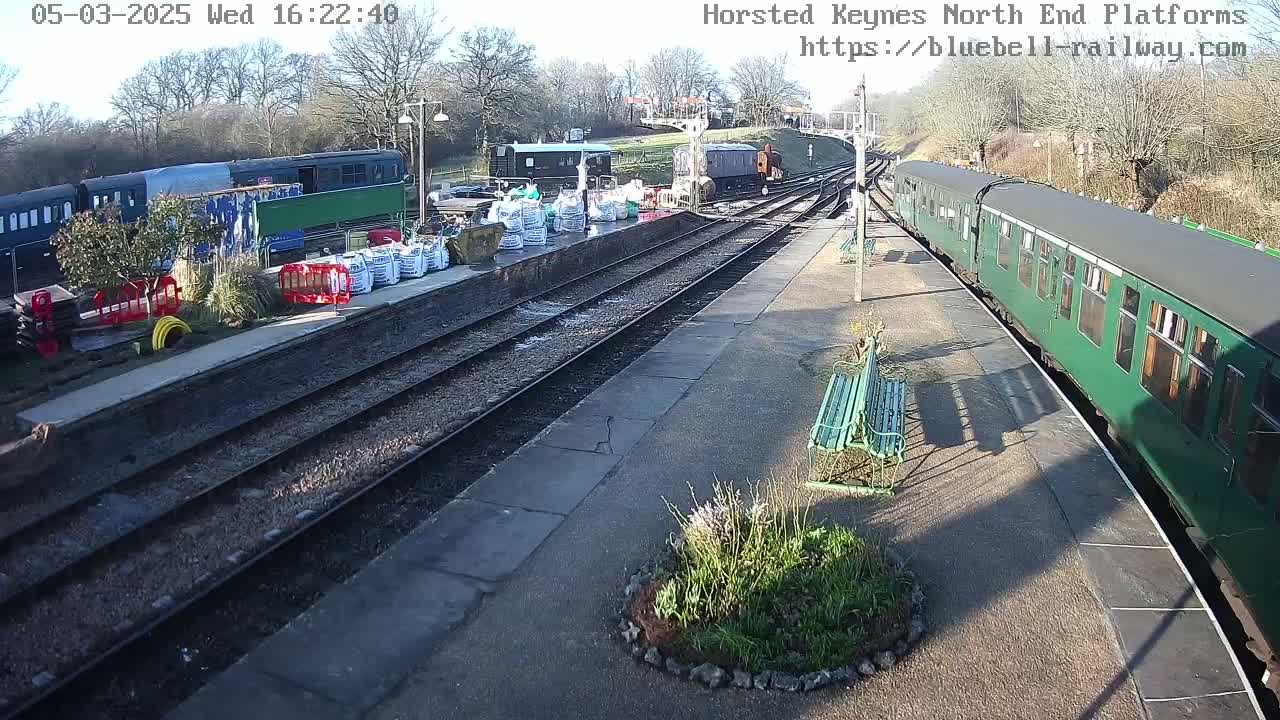 A sunny train station with multiple train cars on the tracks, some construction materials on the platform, and a few benches.
