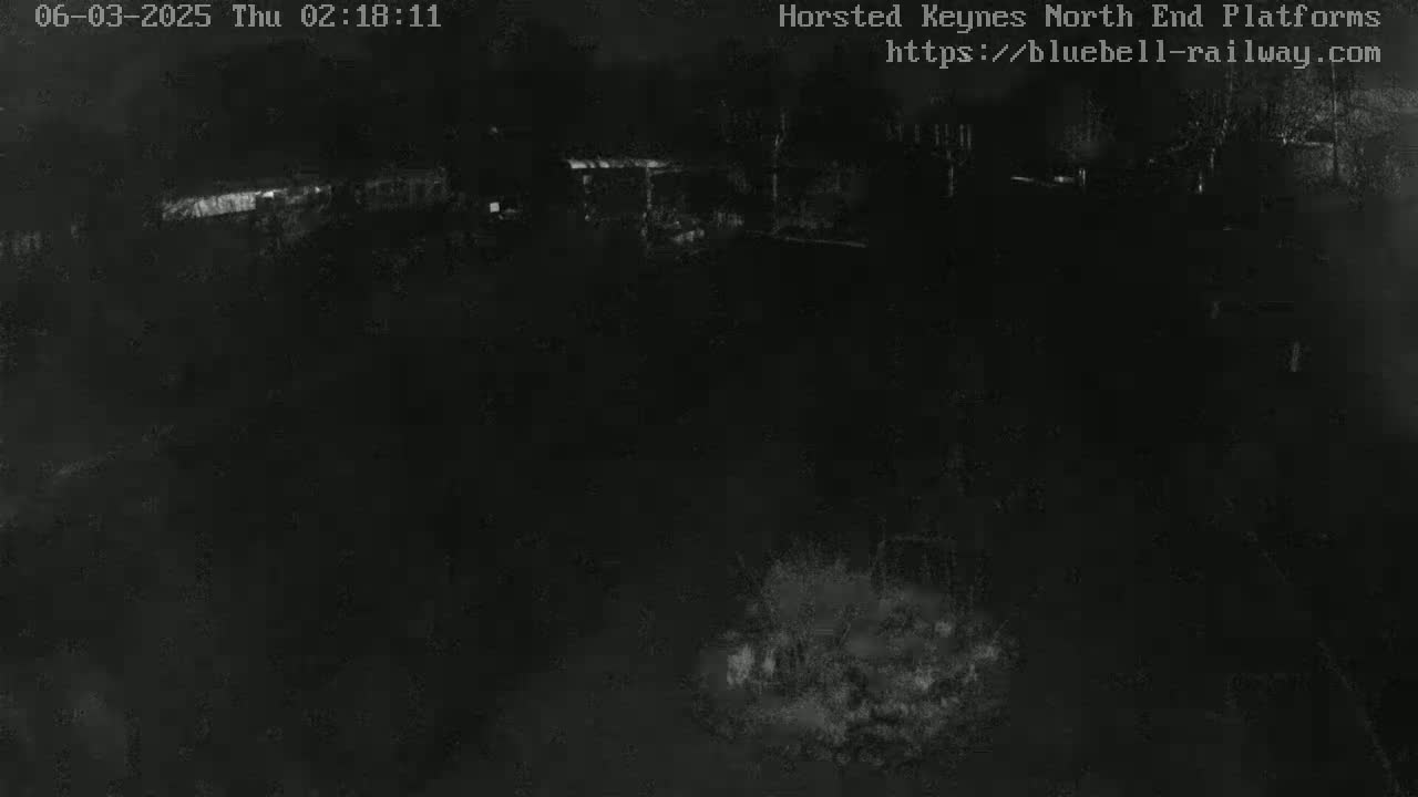 A nighttime, low-light image shows a dark, mostly indistinct outdoor scene with some faintly visible structures and vegetation.