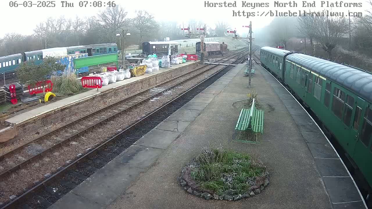 A train station on an overcast day shows several railway carriages, some parked and one departing, along with construction materials and barriers.