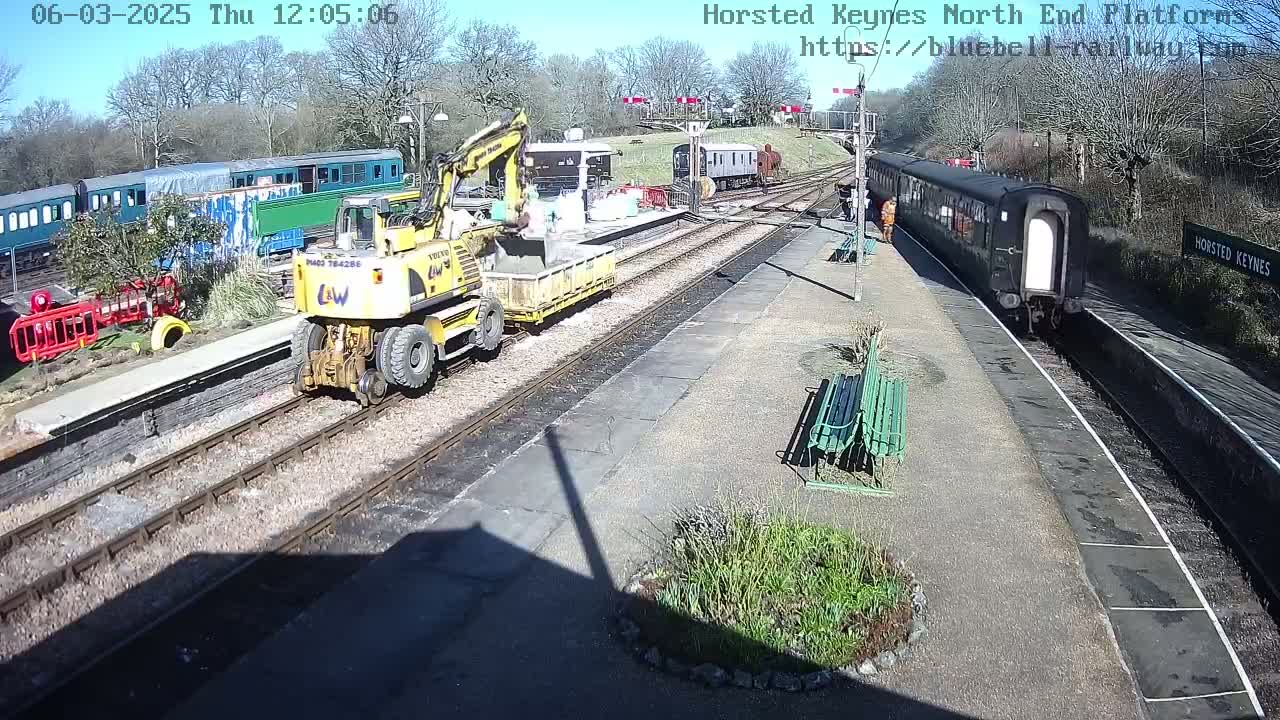 A sunny day at a train station shows a yellow construction vehicle on the tracks near several passenger cars, one of which is departing.