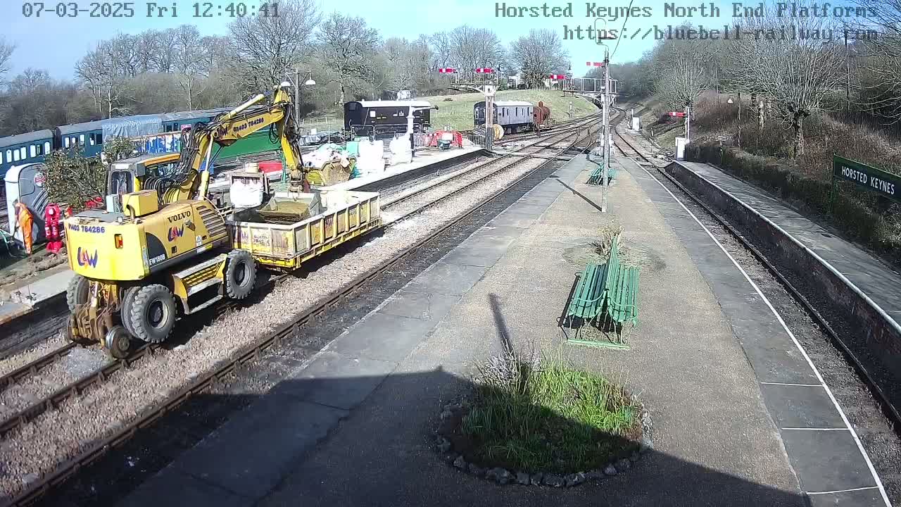 A yellow excavator with a trailer is on a railway platform on a sunny day, near several train cars and some benches.
