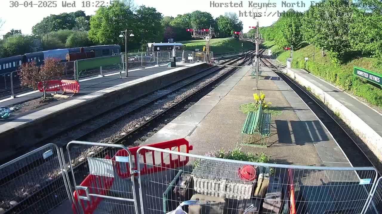 A mostly sunny train station with a passenger train visible in the background, temporary fencing and construction materials scattered around, and several empty benches along the platform.