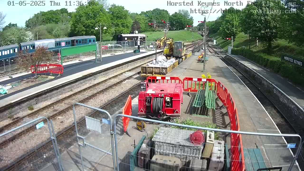 A train station on a sunny day undergoing construction, with railway tracks, trains, construction equipment, and temporary fencing present.