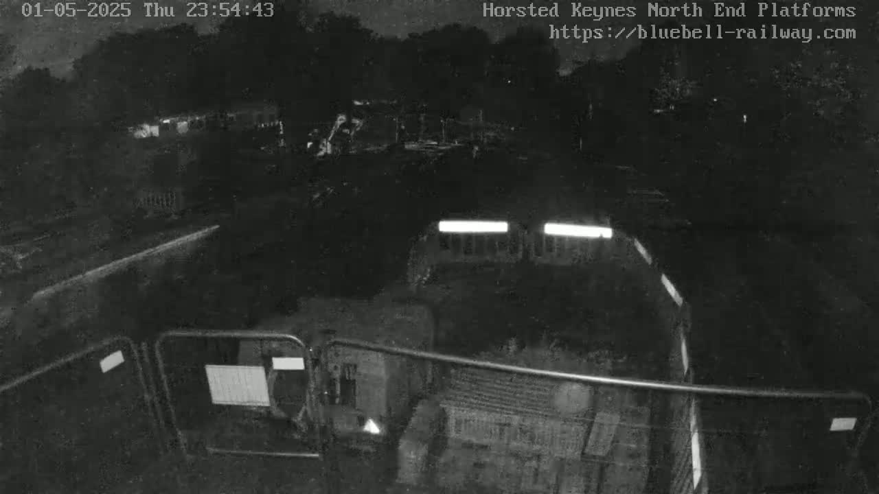 A nighttime, low-light image shows a dark vehicle behind a metal fence in an otherwise empty, dark area.