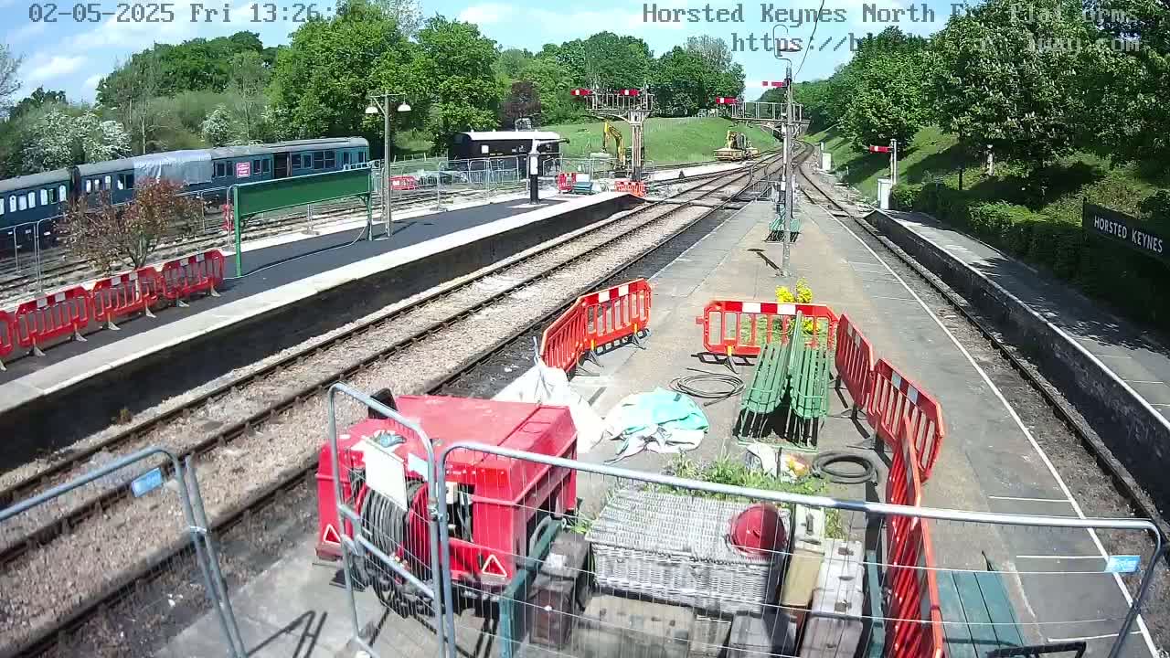 A train station platform under sunny skies shows ongoing construction or maintenance work, with a railway carriage, construction equipment, and temporary fencing.
