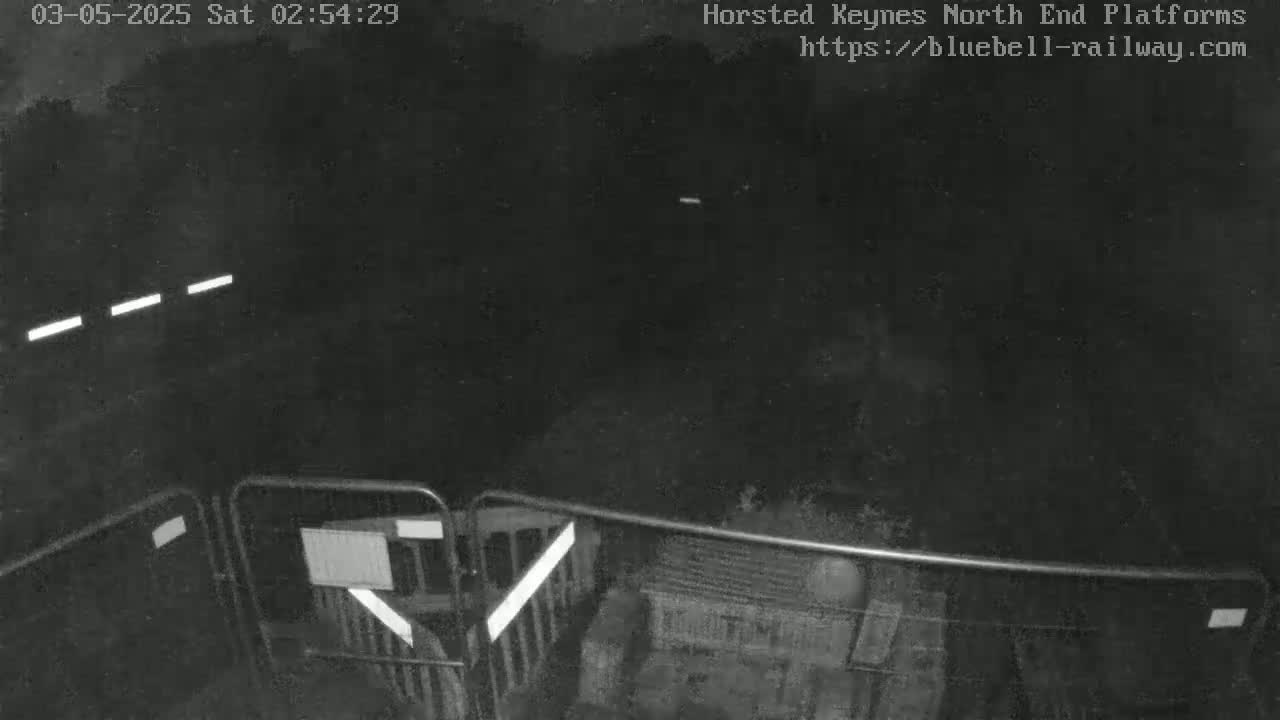 A nighttime, low-light view shows a dark, empty platform area enclosed by metal fencing.