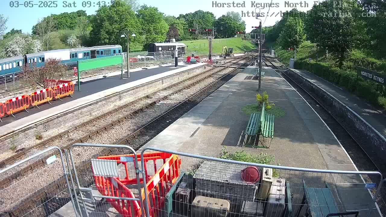 A sunny day at a train station shows railway tracks, a train, construction equipment, and temporary fencing.