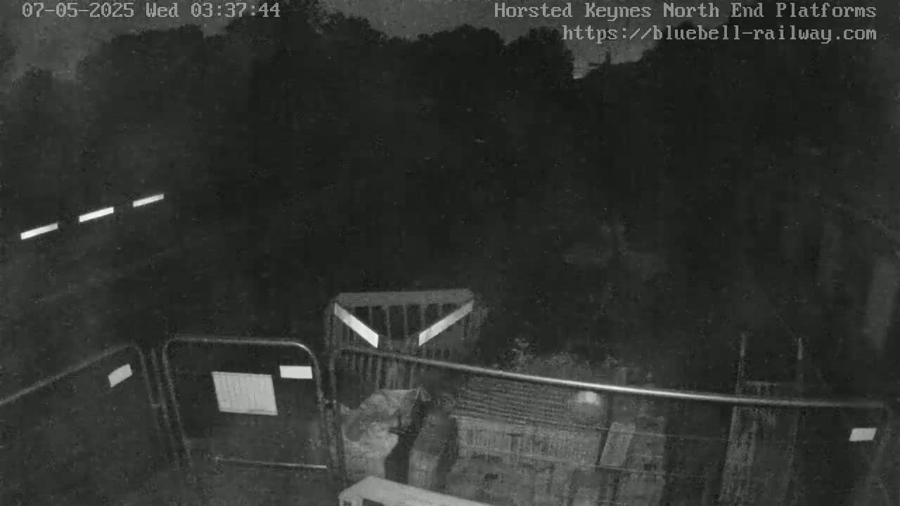 A nighttime, low-light view shows a dark area with metal barriers and fencing, partially obscured by shadows and vegetation.