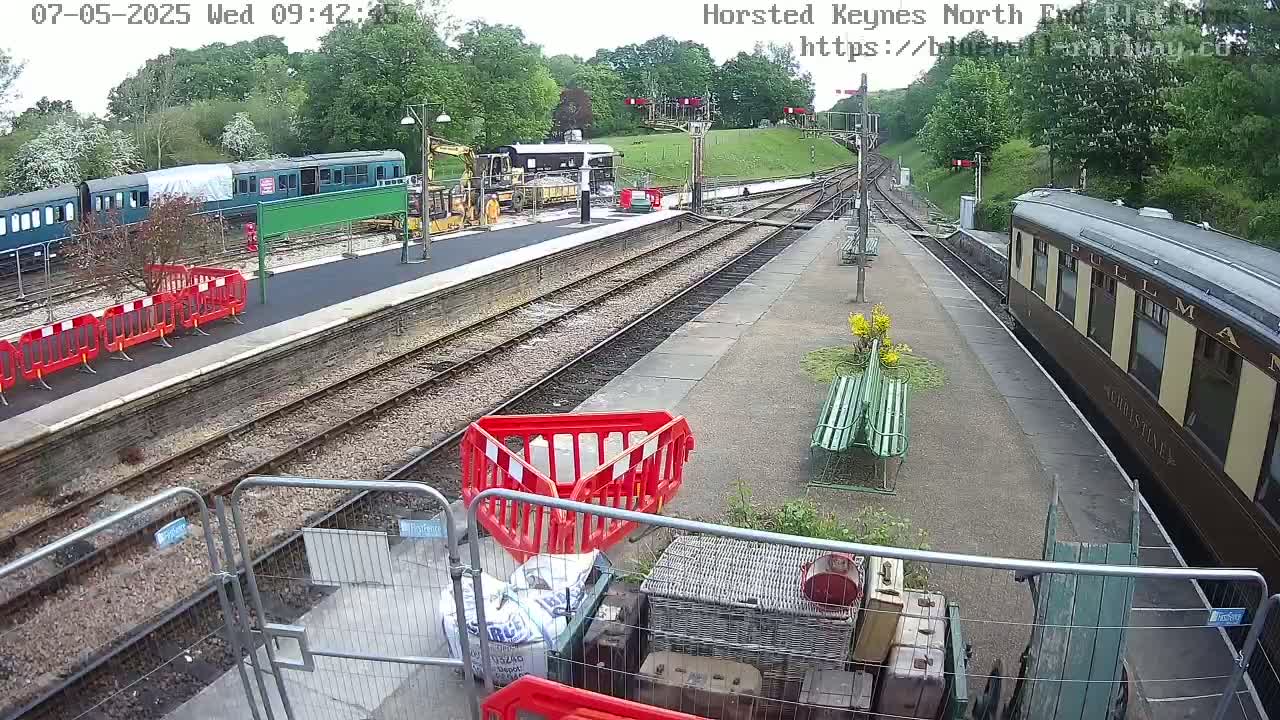 A railway station platform under a partly sunny sky shows several train cars, construction equipment, and temporary safety barriers.