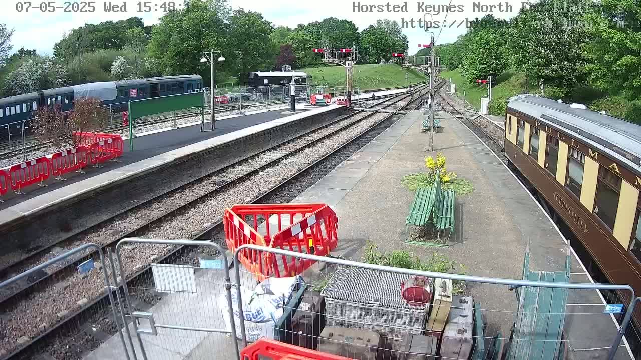 A sunny day at a train station shows several train cars, some construction barriers, and some benches along the platform.