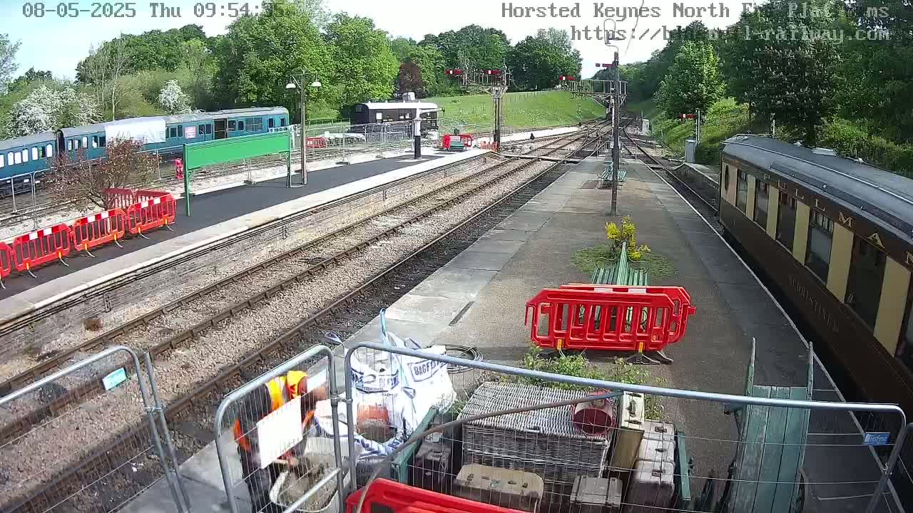 A train station on a sunny day with several train cars, railway tracks, and construction materials and barriers scattered around the platform.