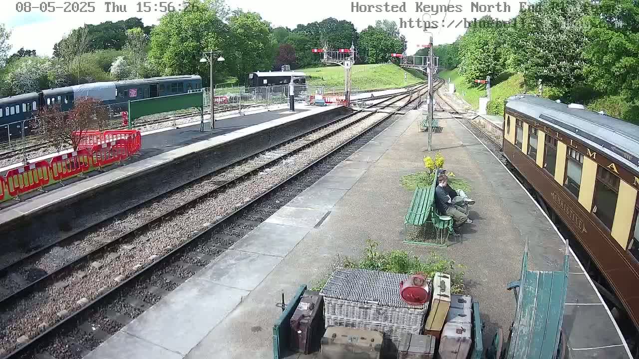 A train station on a sunny day with two passenger trains, one dark green and one brown, with two people sitting on a bench.
