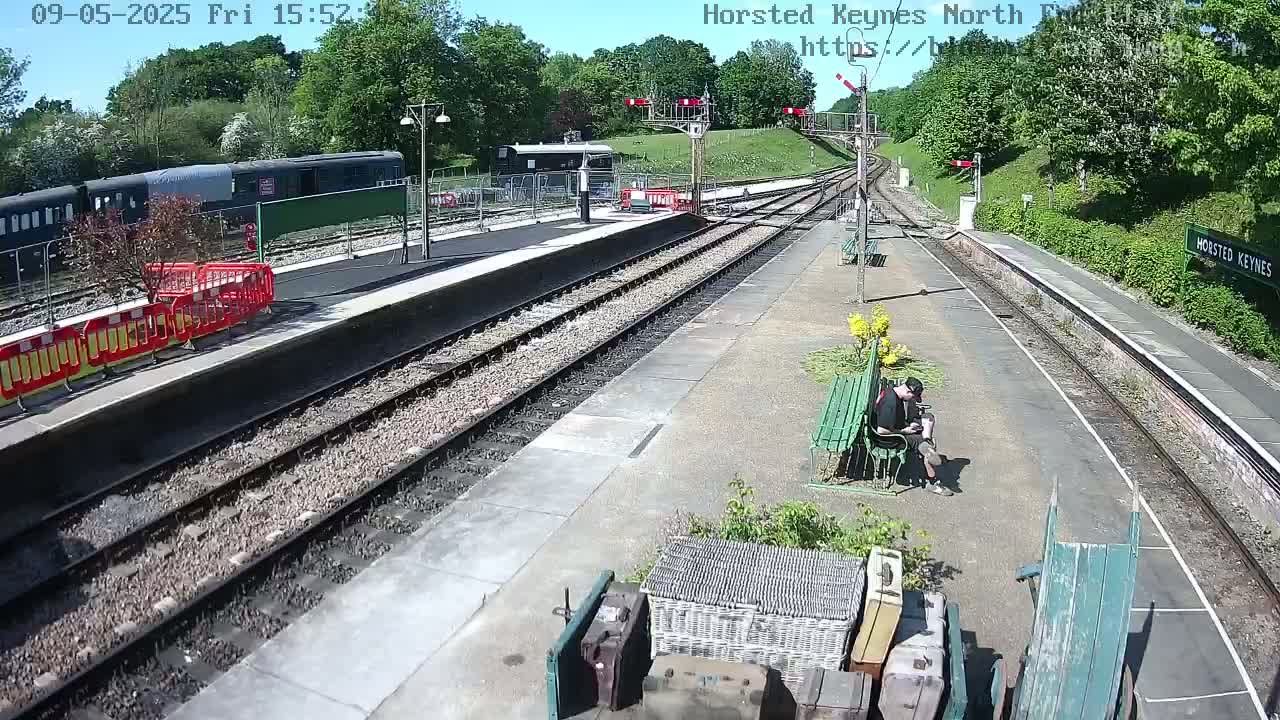 A sunny view of a train station platform with a person sitting on a bench, railway tracks, and a train parked nearby.