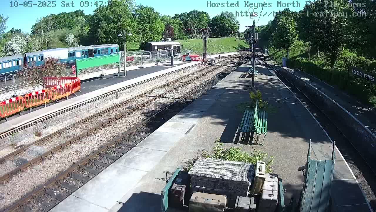 A sunny day at a train station with a train parked on the tracks, maintenance equipment on the platform, and a few benches.