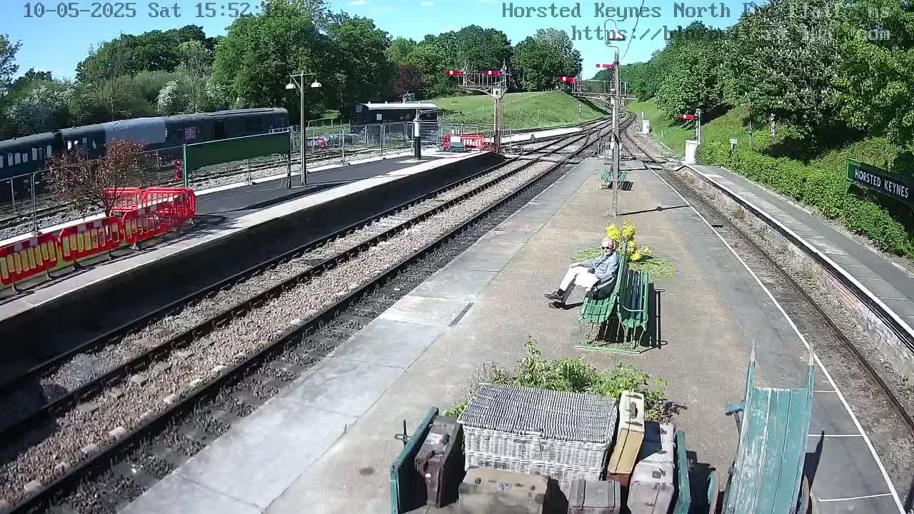 A sunny day at a train station shows multiple train tracks, a train in the distance, a person sitting on a bench, and various pieces of luggage and a wheelbarrow.