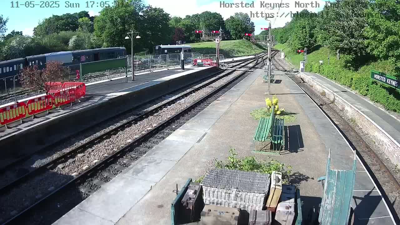 A sunny day at a train station with multiple train tracks, a dark-colored train parked nearby, and several benches and gardening supplies on the platform.