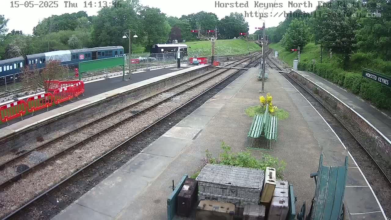 A train station platform with multiple train tracks, a partially covered train, and some luggage on a cart, under a partly sunny sky.
