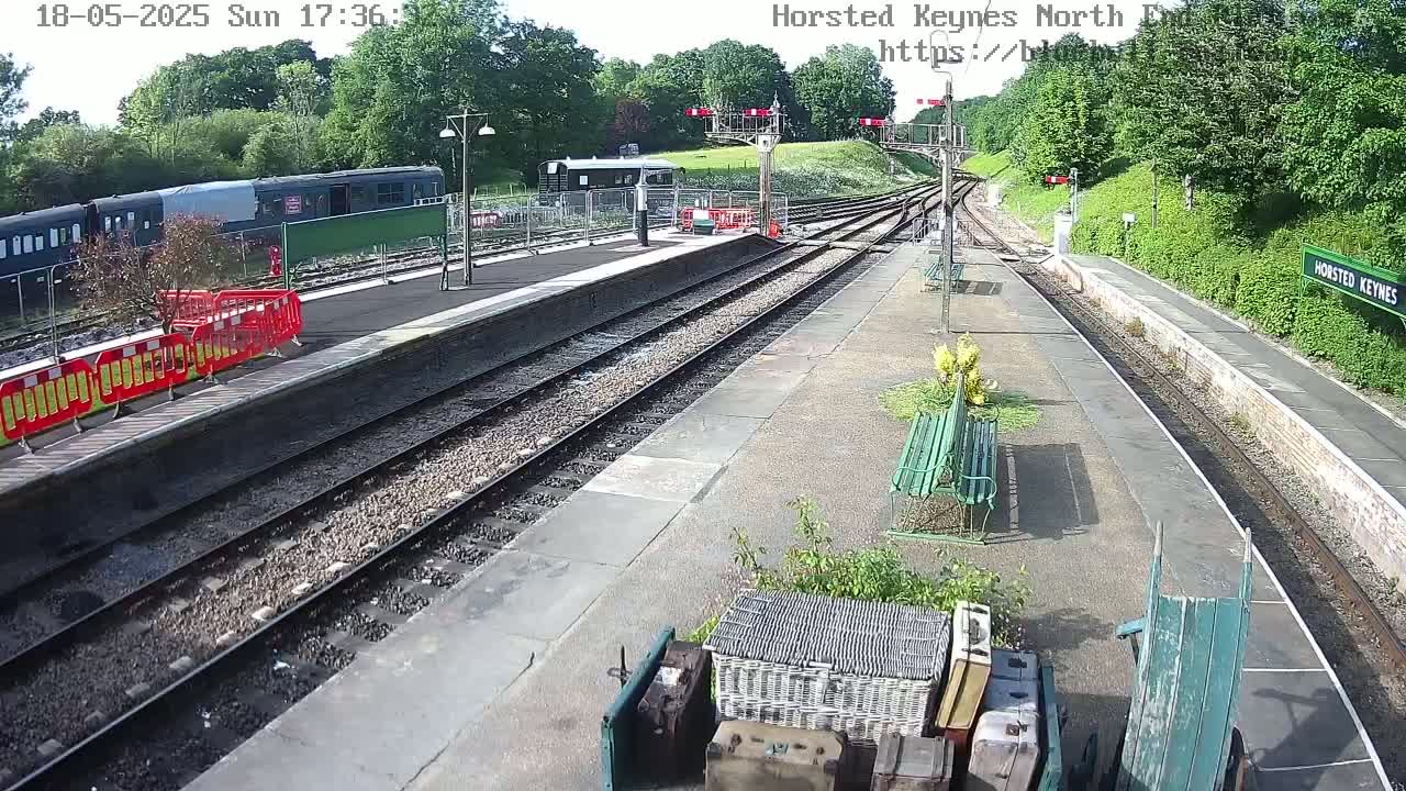 A train station platform with multiple train tracks, a partially covered train, and some luggage on a wheelbarrow, on a sunny day.