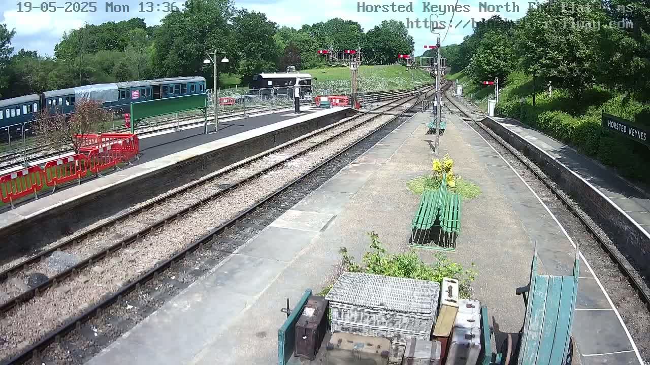 A train station platform on a sunny day with train cars visible, several sets of train tracks, and some luggage near a green hand truck.