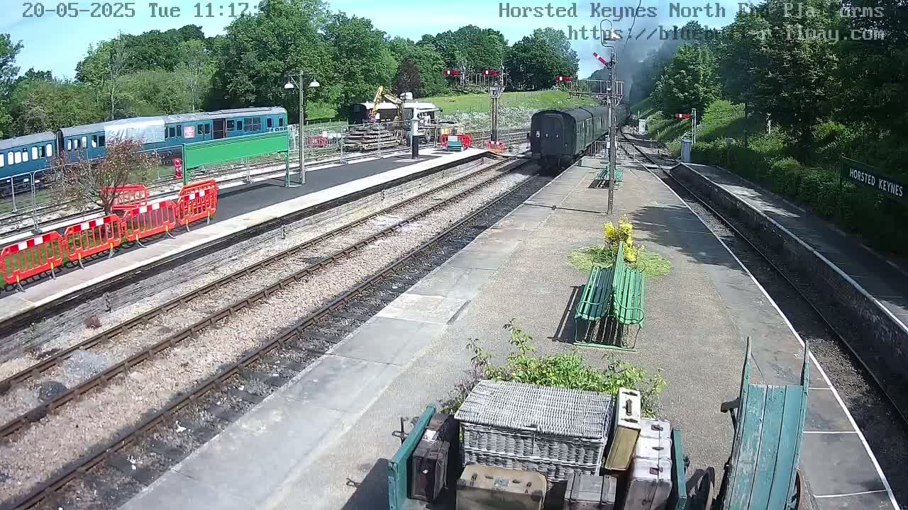 A sunny day at a train station shows a dark-colored train departing and a teal train at the platform, with construction equipment visible nearby.