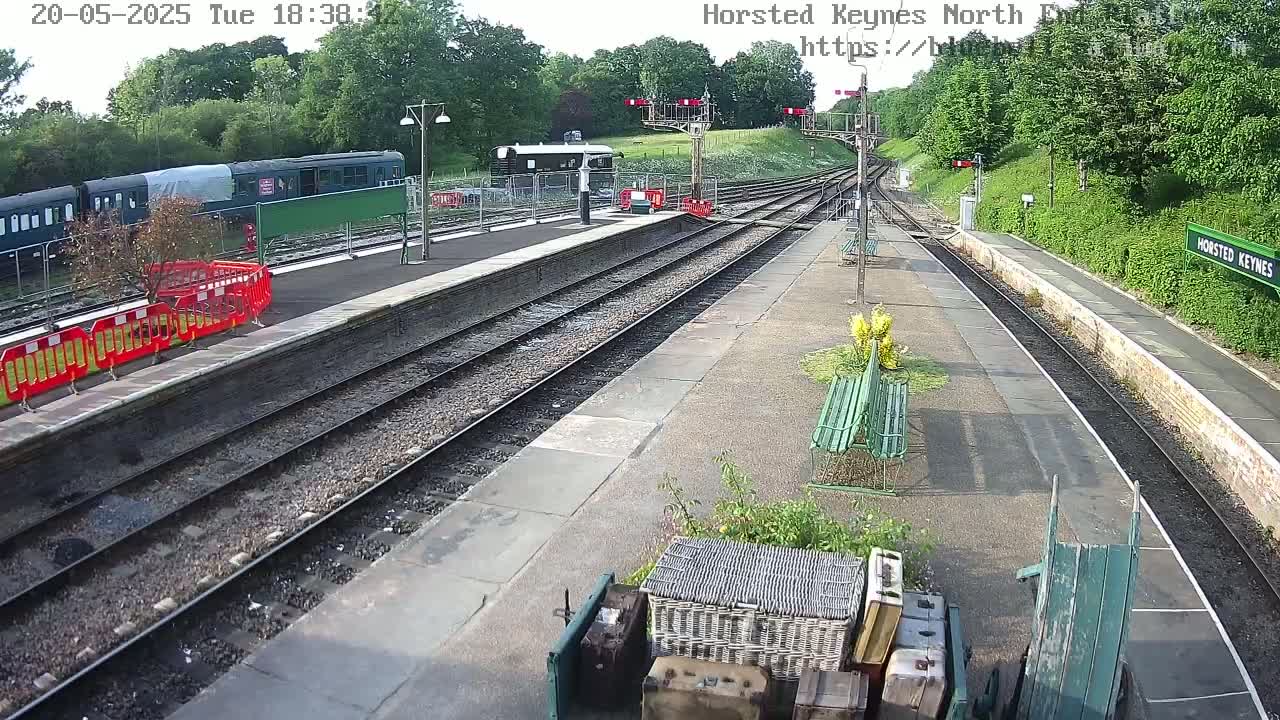 A train station platform with multiple train tracks, a partially covered train car, and several benches, under a partly sunny sky.