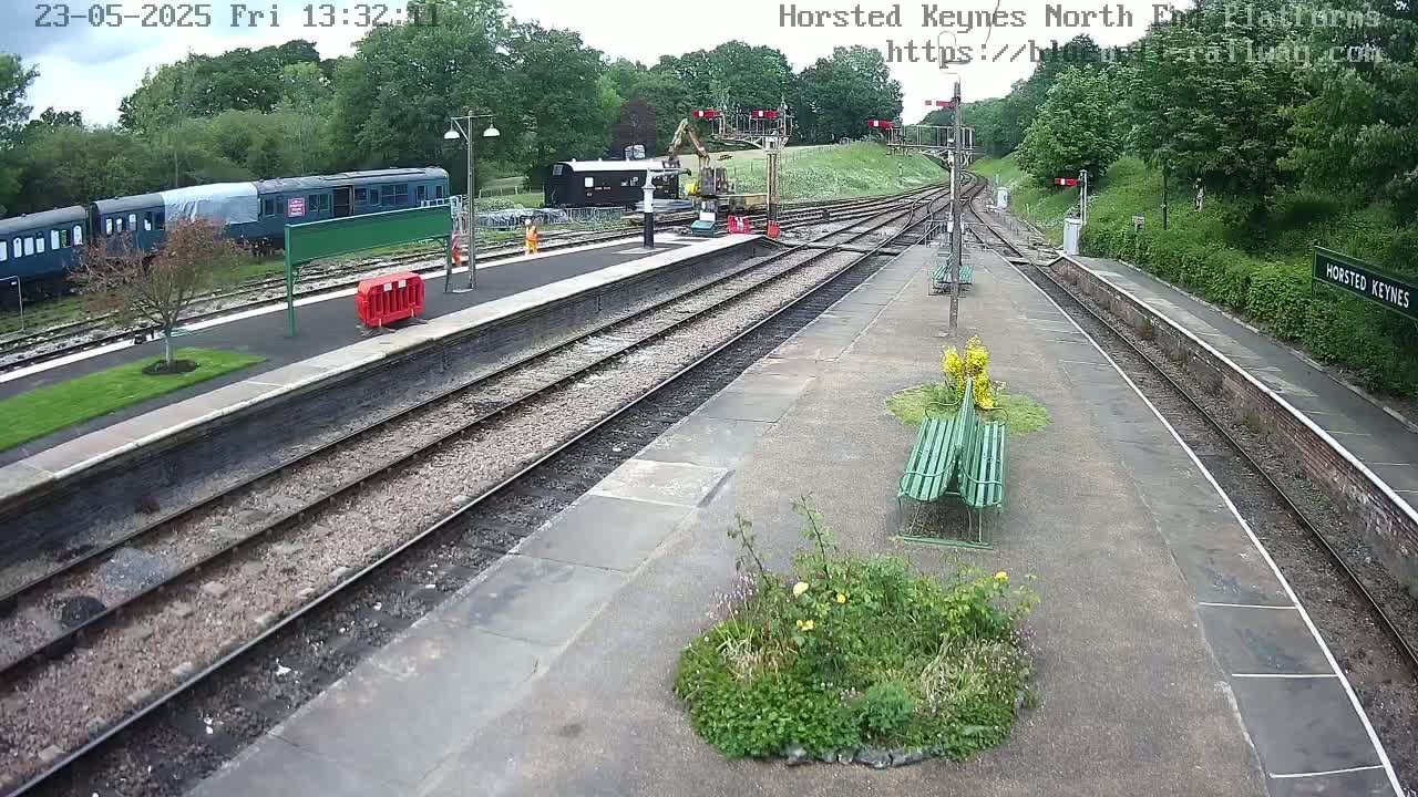 A railway station platform with several train carriages, construction equipment, and greenery under partly cloudy skies.