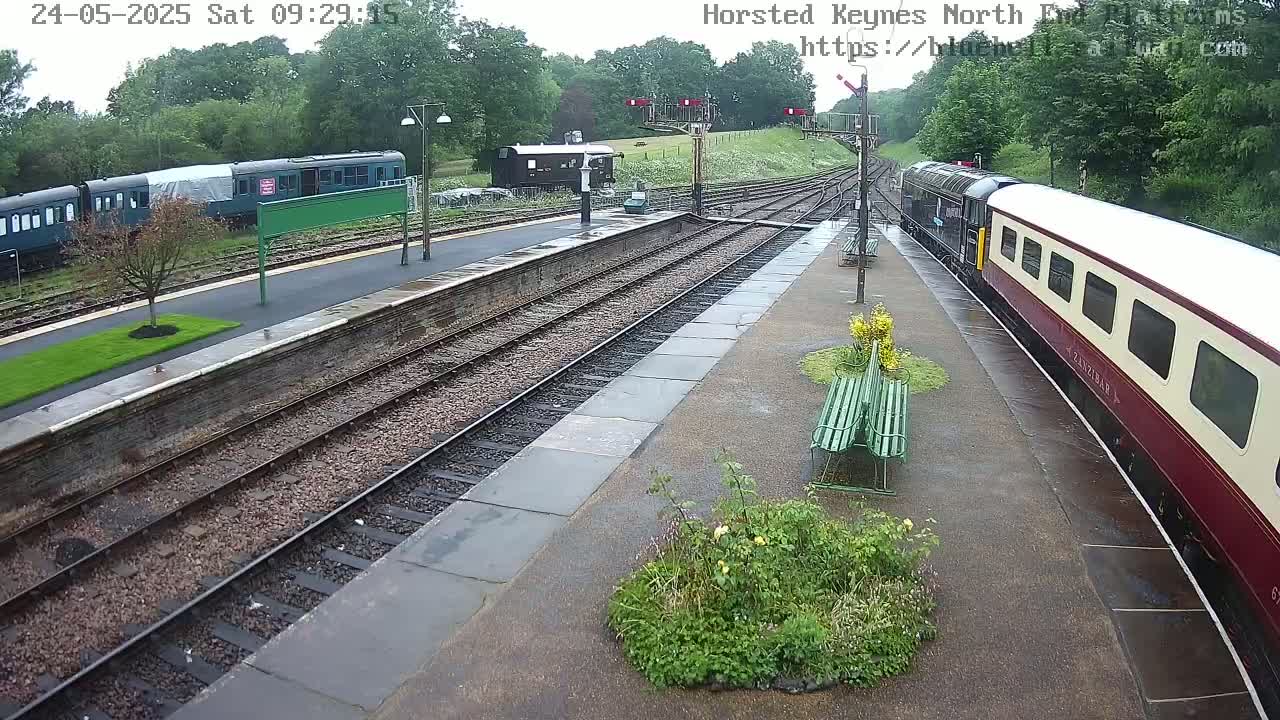 A train pulls into a rain-slicked railway station platform where other trains and railway buildings are visible.
