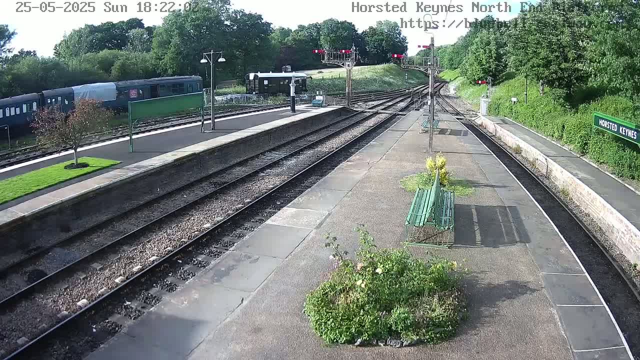 A mostly sunny view of a train station platform with a train parked nearby, surrounded by green trees and bushes.