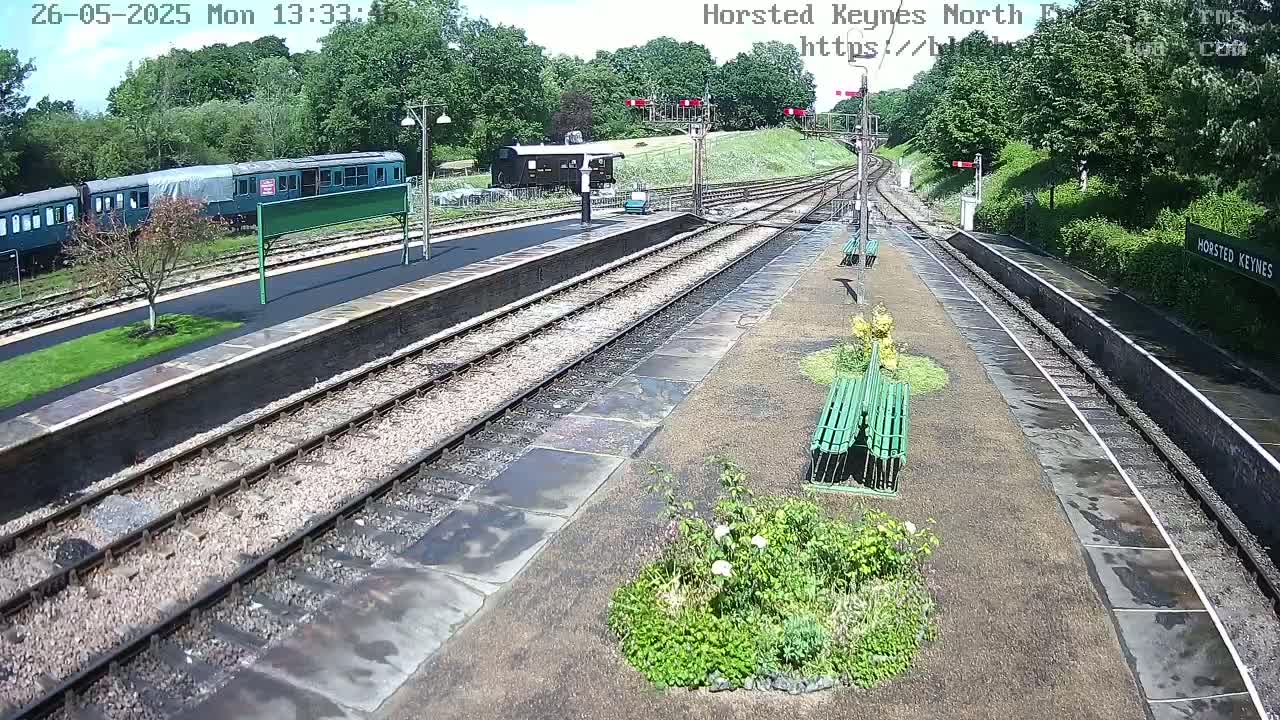 A train station platform with a dark-green train and several benches under a partly sunny sky.