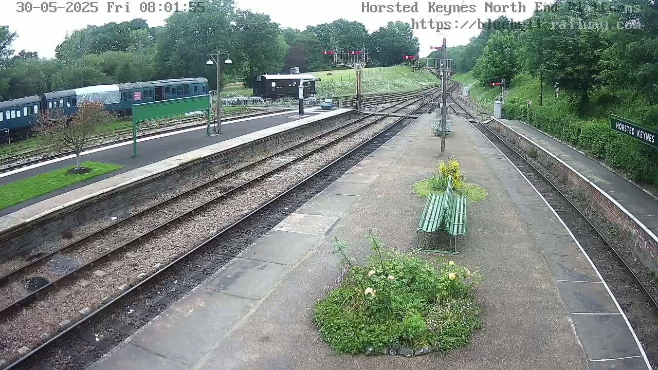 A mostly sunny view of a train station platform with several train cars and a few benches, surrounded by greenery.