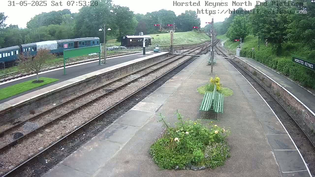 A train station platform with multiple train tracks, railway cars, and benches, surrounded by greenery under an overcast sky.