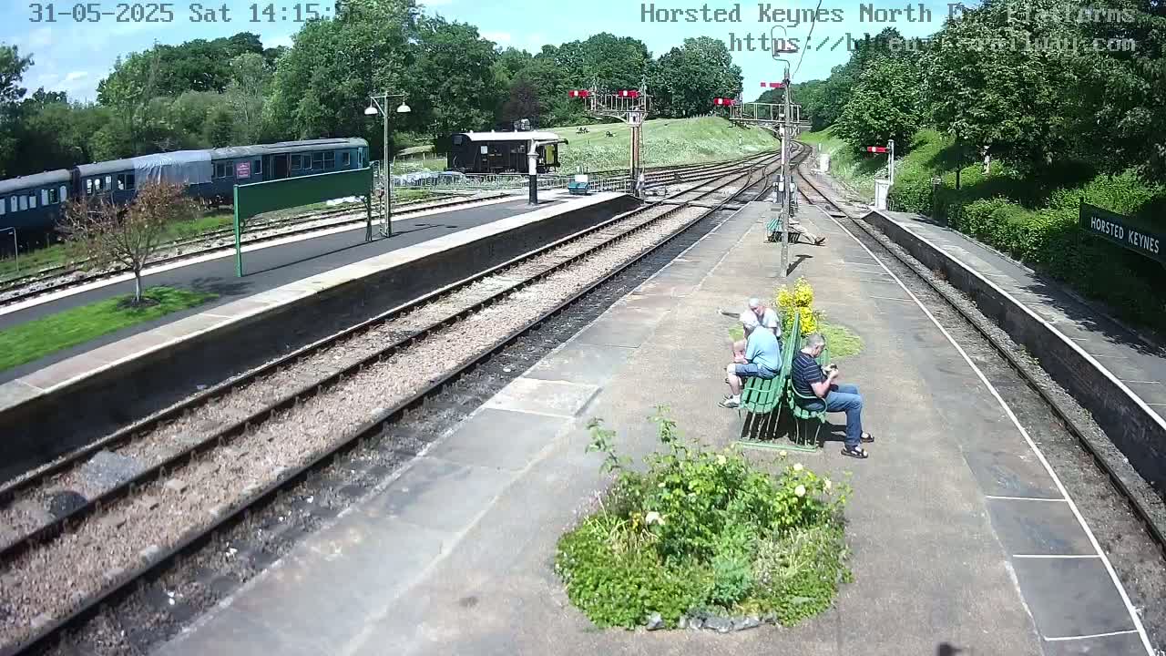 A sunny day at a train station with a dark green train parked on a side track, three people sitting on a bench, and multiple train tracks leading to the horizon.