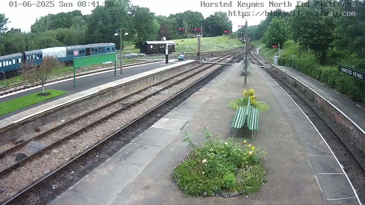 A mostly sunny view of a train station platform with several railway tracks, a parked train, and some greenery.