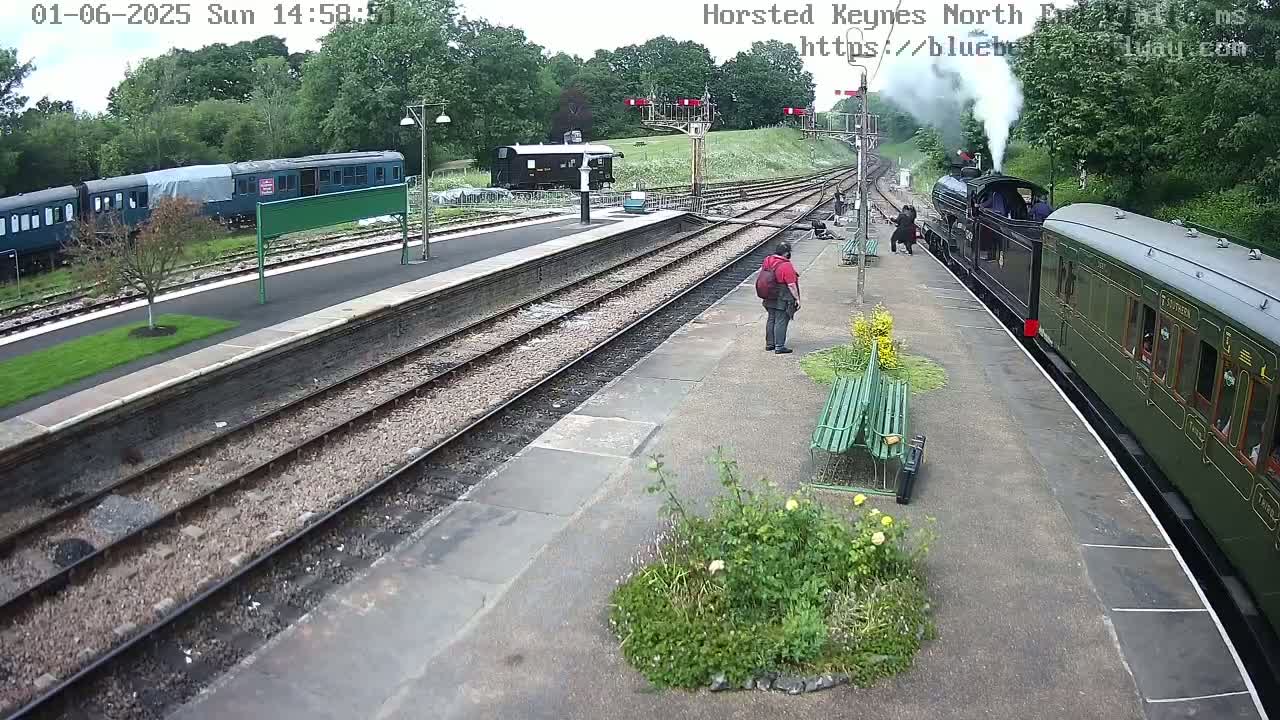 A steam train is arriving at a station platform on a partly sunny day, with a few people standing nearby and other trains parked on adjacent tracks.