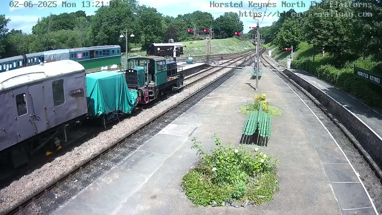 A train station on a sunny day with several train cars and a diesel locomotive on the tracks, and green benches along the platform.