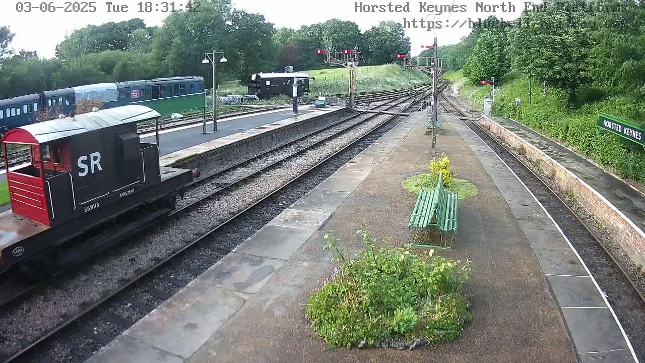 A railway station platform with a red and black railway carriage and several train cars visible, under partly cloudy skies.
