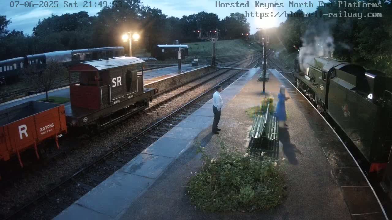 A nighttime scene at a train station shows a steam train arriving at a platform, where two people stand near a bench, and several railcars are parked nearby.