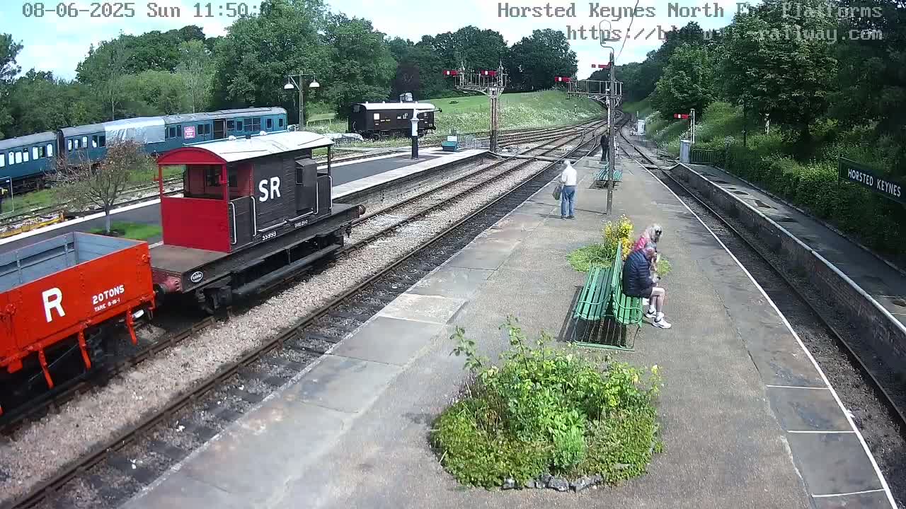 A railway station platform on a sunny day with several train cars, some people sitting on a bench, and various railway equipment.