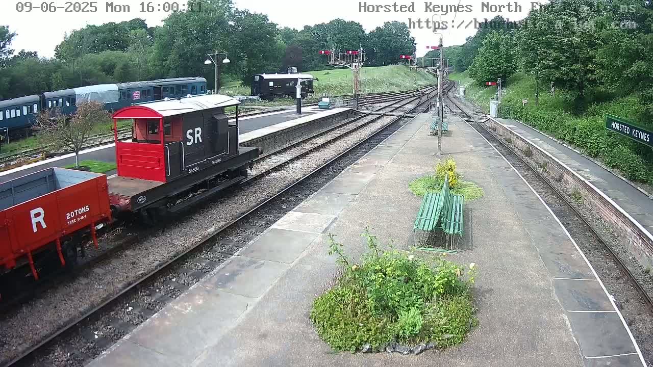 A railway station platform with several train cars, including a red and black locomotive, under partly cloudy skies.