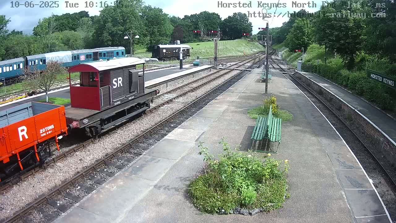 A railway station platform on a sunny day with several railway cars, including a red boxcar and a dark-colored passenger car, visible along the tracks.