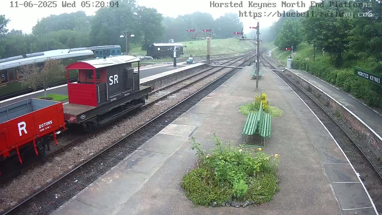 A railway platform on an overcast day shows a red and black railway carriage coupled to a red railway wagon, alongside a longer train; green benches and vegetation are present on the platform.