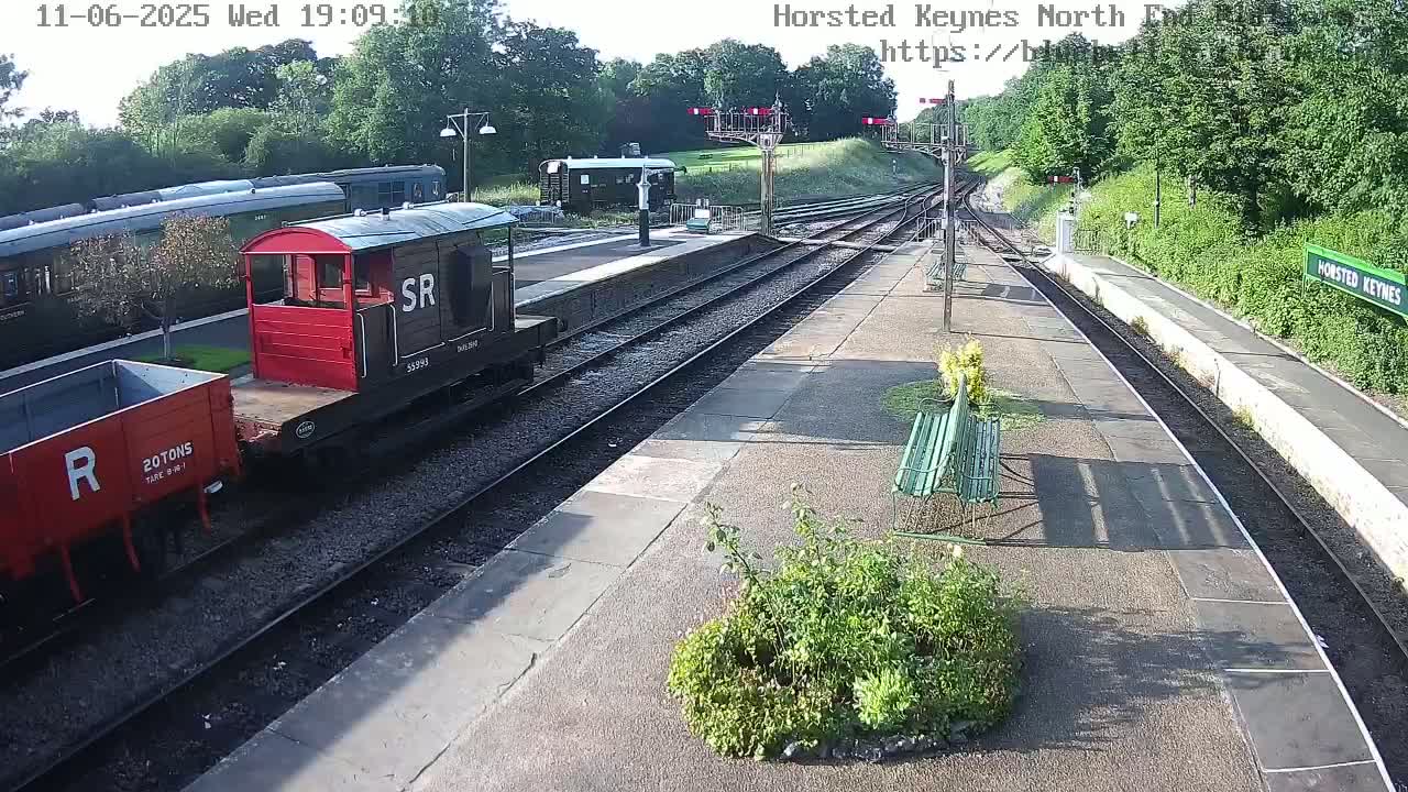 A railway station platform on a sunny day with a red railway carriage, a dark-colored train, and a red wagon on the tracks.
