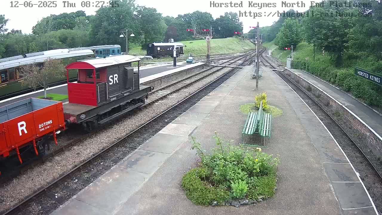 A train consisting of a red and black engine, a red boxcar, and a long green passenger car is parked at a railway station on an overcast day.