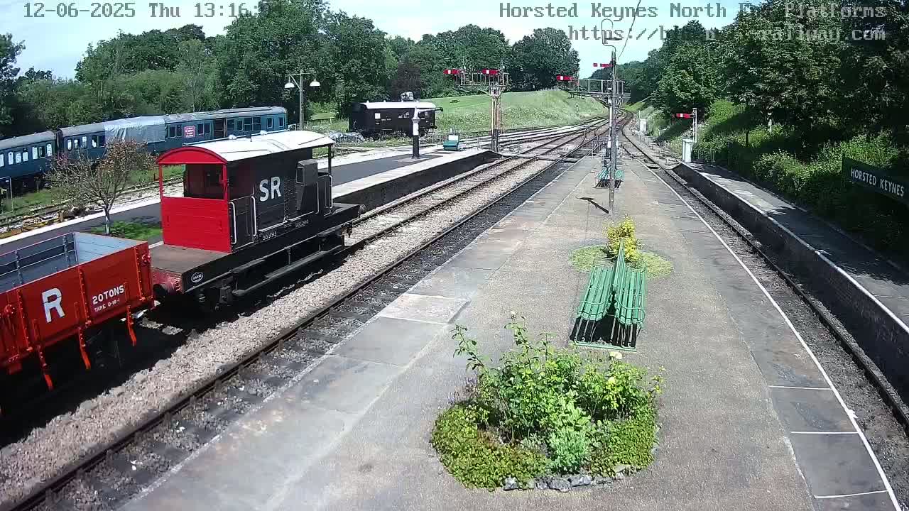 A railway station platform on a sunny day with a small red and black train and a red railway wagon on the tracks.