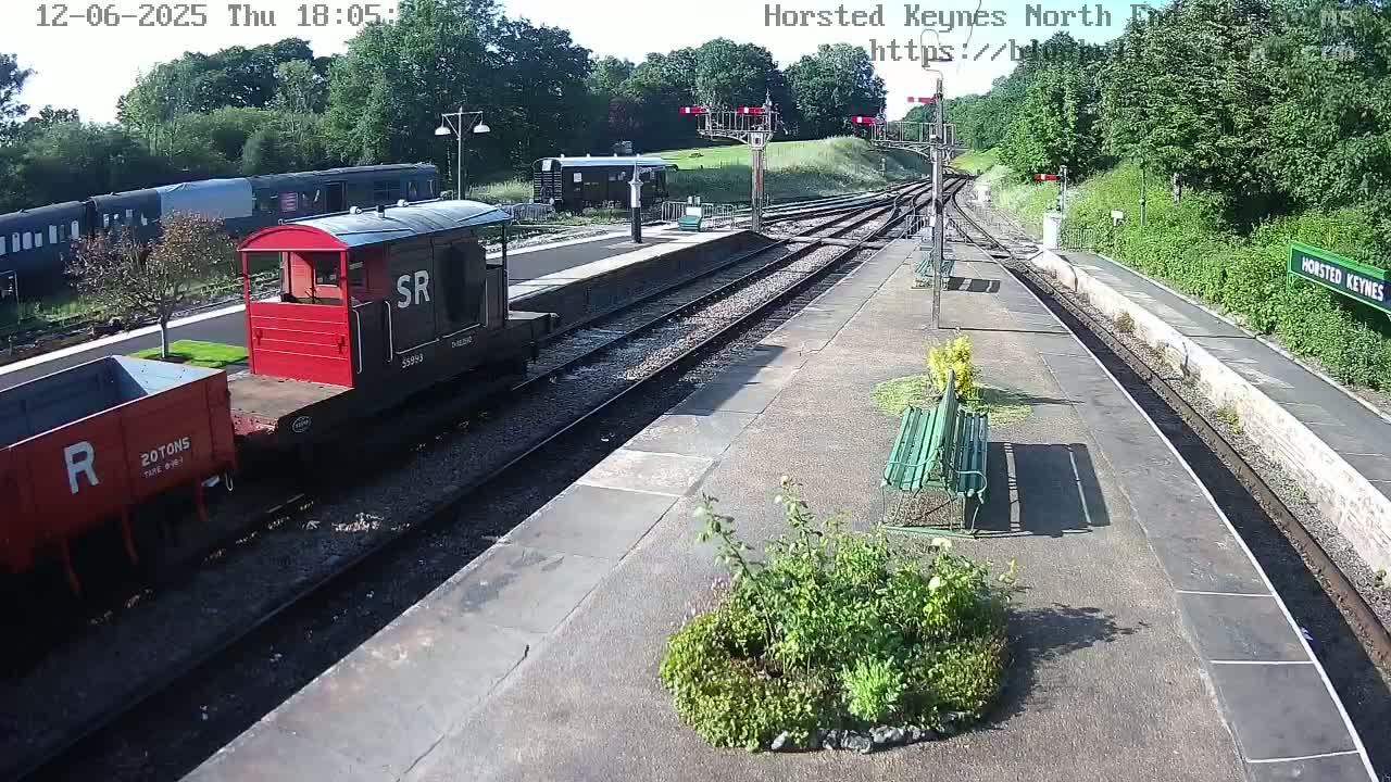 A railway station platform on a sunny day with several train cars, including a red one, and a small garden bed.