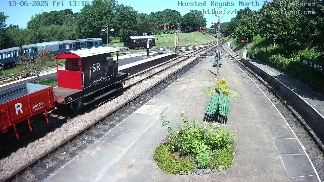 A red and black railway engine and a red railway car are parked on a train track at a station on a sunny day.
