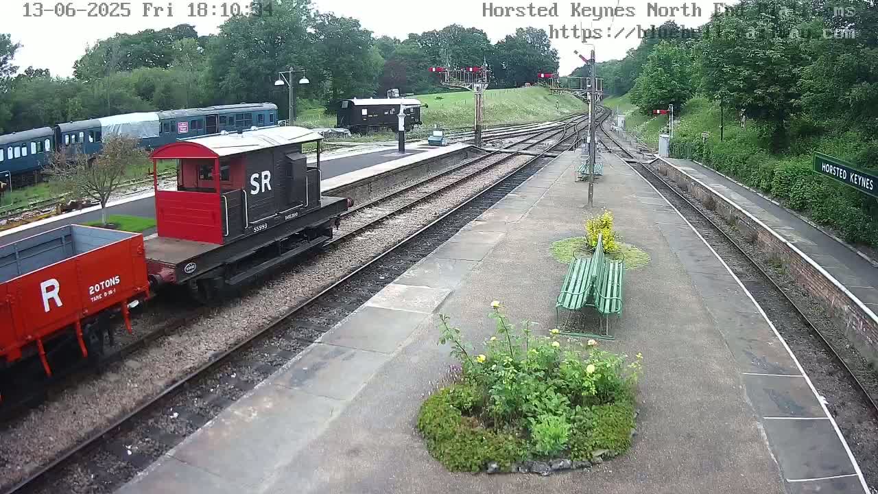A train station on an overcast day with railway tracks, a red and black railway carriage, and a red freight car, surrounded by greenery and benches.