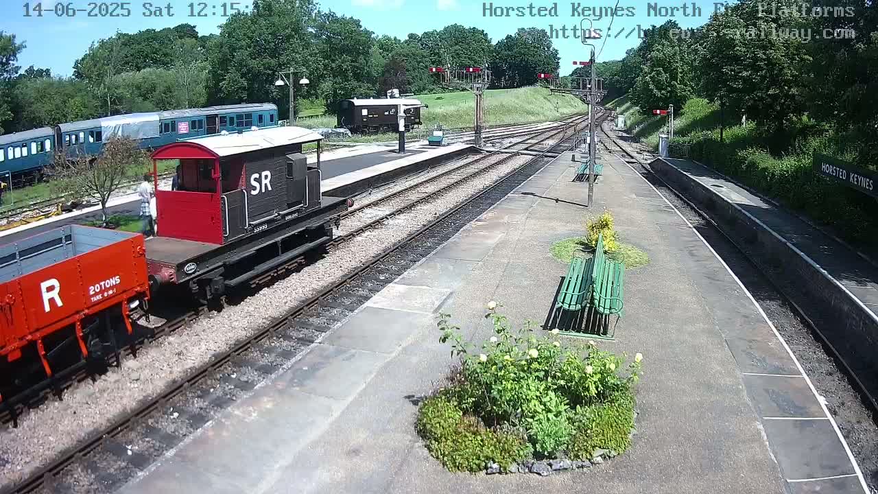 A sunny day at a train station shows several railway cars, including a red boxcar and a dark-colored engine, parked along the platform with green benches and a small garden.